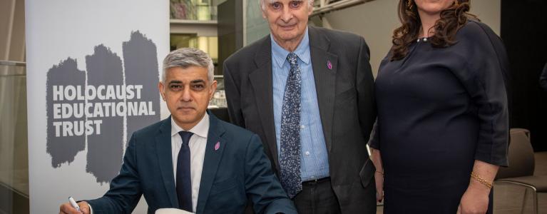 Sadiq Khan seated signing a book of commitment with Karen Pollock stood next to him