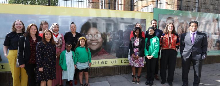 School children and artists and head teacher stand on pavement in front of mural outside school featuring Doctor Beryl Gilroy