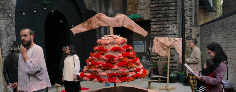 A sculpture of a dress with red frills stands on a pedestal, people wander around it looking at the exhibition