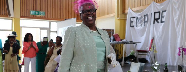 An older black woman walks proudly to camera, smiling, wearing a silk green dress, jacket, with lace gloves, and pink hair fascinator