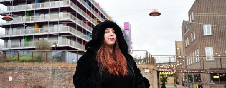 A young white woman wearing a large black hooded jacket, she looks in the distance, a backdrop of buildings and urban bridges and structures