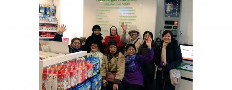 a group of older people strike a pose in a pharmacy, they wear scarves and coats, wave and smile to camera.