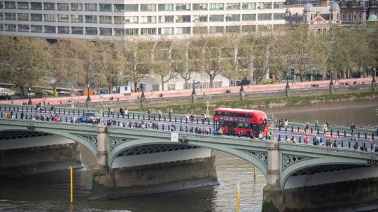 A red bus driving over Westminster bridge with the Thames river, members of the public and trees visible