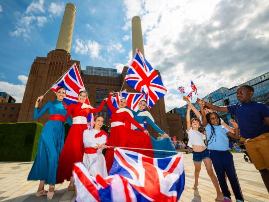A group of women dressed in red, blue and white holding Union Jack flags alongside a group of children waving smaller flags, in front of Battersea Power Station