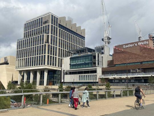 A group of people walking and a cyclist cross a bridge in front of four distinct modern buildings in the final phase of construction with two large cranes behind the buildings.