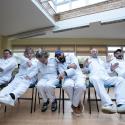 6 people sit in a row, wearing white boiler suits, dynamically waving, pointing and making faces playfully