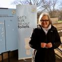 Caroline Russell poses with a smile next to a banner with lots of yes votes for more public toilets on the tube network.