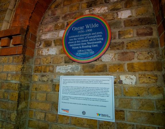 A blue plaque with a rainbow edge mounted on a yellow brick wall. The plaque is dedicated to OScar WIlde and reads 'Oscar Wilde 1854-1900, celebrated playwright and poet, 