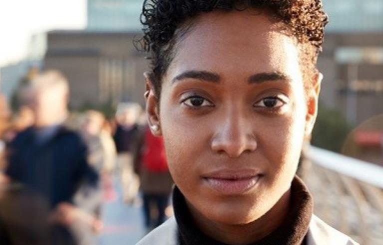 Young black woman stands looking into the camera on millenium bridge in London as people pass her by.