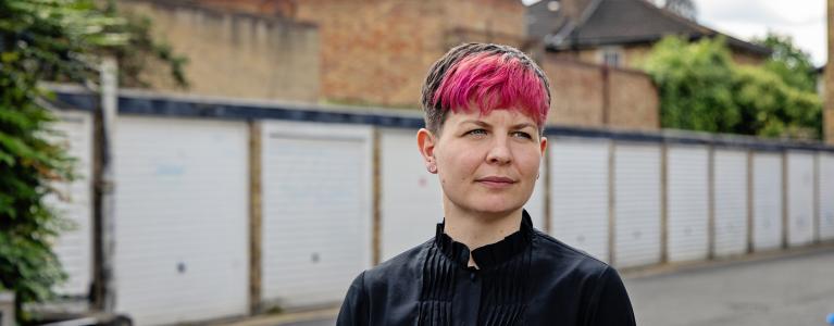 Zoë Garbett stands in a London alley in front of garages. The garages are painted white. Zoë is unsmiling. 