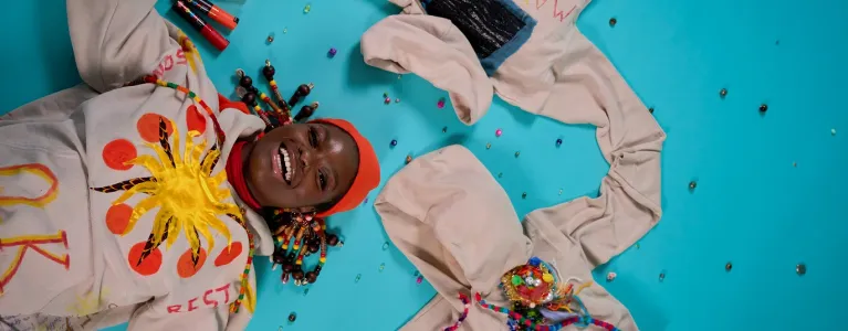 A young person with colourful braids lies on the floor looking straight up at the camera with a wide smile, wearing a hoodie with a sun that says ‘OK’ and holding two other hoodies with colourful writing and beads spread on the floor. It is vibrant and colourful.