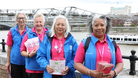 Four Team London Ambassadors volunteers posing for picture in front of River Thames and holding Let's Do London campaign leaflets