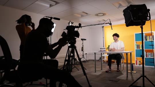 Behind the scenes of filming; a camera and its handler filming a presenter sat at a desk with lighting and mic visible.
