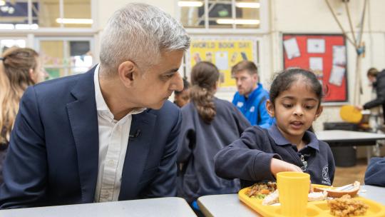 Mayor of London, Sadiq Khan, sitting beside a primary school student in a school canteen.