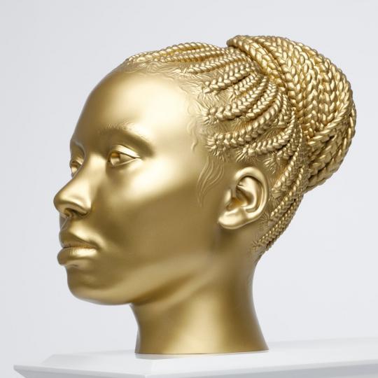 Close up view of the head of a black woman with braided hair in a bun, in a shiny gold colour, on a light grey plinth angled