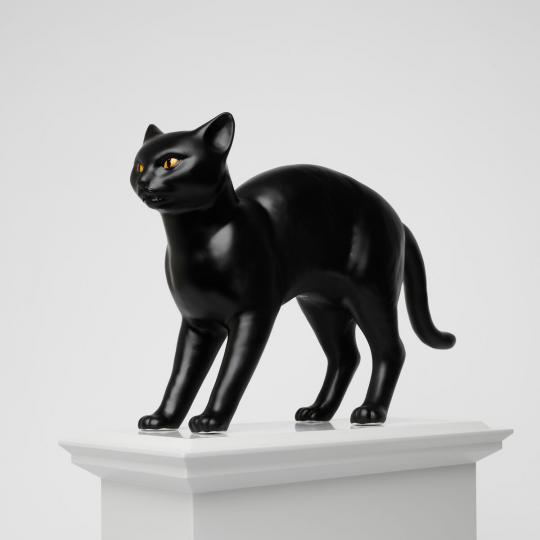 Side angled view of black ceramic cat, with orange eyes, and an arched back, on a light grey plinth