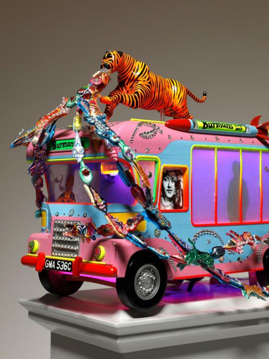 Close up side angle view of blue and pink tone ice cream van with a tiger on top with jewels streaming out of its mouth on a light grey plinth