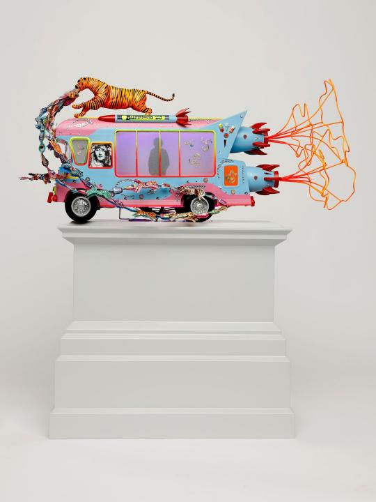 Front view of Blue and pink tone ice cream van with a tiger on top with jewels streaming out of its mouth, a silhouette of a driver in the front window, on a light grey plinth