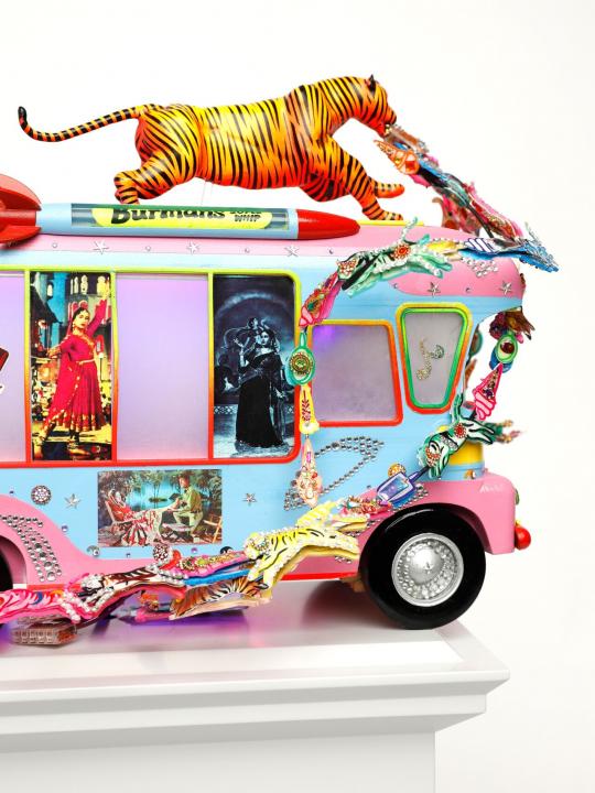 Close up side view of blue and pink tone ice cream van with a tiger on top with jewels streaming out of its mouth, pictures of Indian figures in the side window, on a light grey plinth