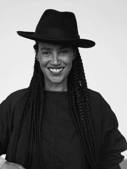 Black and white portrait of a woman smiling, with long braided hair, wearing black clothing and a large black hat.