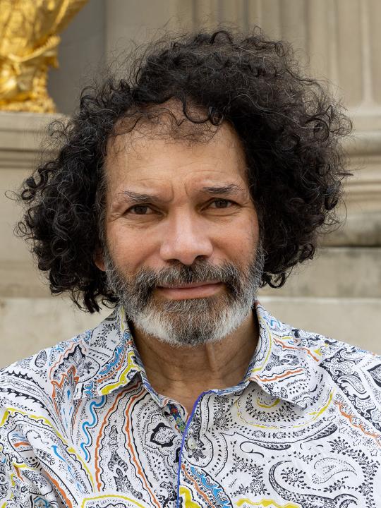 A photograph portrait of a man, with dark curly hair and greying beard, wearing a patterned shirt.