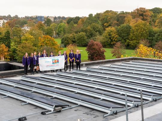 Students holding a banner on top of school roof beside solar power generators. 