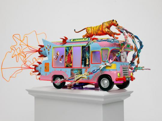 Side view of Blue and pink tone ice cream van with a tiger on top with jewels streaming out of its mouth, pictures of Indian figures on the side window and orange exhaust fumes, on a light grey plinth