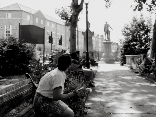 Person sits on a bench listening to their phone while looking at a statue of William Gladstone