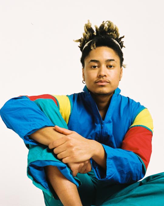 A picture of the artist Khaleb Brooks. He is wearing a brightly-coloured jacket, and sitting with his hands clasped over his right knee. He is mixed-race and has a single earring in his right ear.