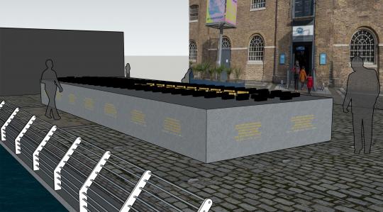 A computer generated image of a streetscape with cobbled street, and a large grey, waist height, solid block, with gold coloured text around the side of the and on top of the grey block are smaller black blocks and a few gold coloured blocks. 