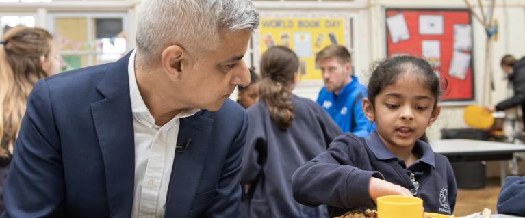 Mayor of London, Sadiq Khan, sitting beside a primary school student in a school canteen.