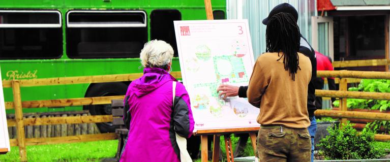 A map in a black cap, and black jumper and blue trousers, largely obscured from view, shows a woman with white hair and a pink coat and jeans and a woman with dreadlocks in a caramel top and camouflage trousers a map on a wooden easel. Behind them is a section of enclosed parkland and behind that is a green bus.