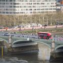 A red bus driving over Westminster bridge with the Thames river, members of the public and trees visible