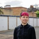 Zoë Garbett stands in a London alley in front of garages. The garages are painted white. Zoë is unsmiling. 